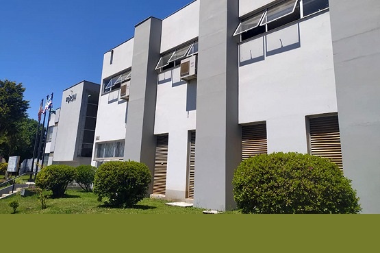 A imagem mostra um prédio de dois andares com uma fachada moderna em tons de cinza claro e branco. A arquitetura inclui janelas retangulares grandes, algumas das quais são protegidas por persianas externas de cor madeira, oferecendo um contraste interessante com a cor predominante do prédio. Na frente, um pequeno jardim bem cuidado com grama verde e alguns arbustos adiciona um toque de natureza ao cenário urbano. Bandeiras estão visíveis, hasteadas em frente ao prédio, indicando possivelmente uma instituição governamental ou corporativa. A imagem transmite uma sensação de ordem e funcionalidade, típica de edifícios administrativos ou empresariais.