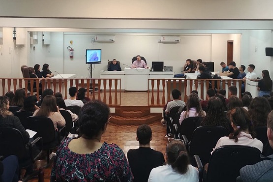 A imagem mostra uma sala de conferências ou tribunal com várias pessoas sentadas. Na parte da frente, há uma mesa comprida com algumas pessoas sentadas atrás dela, possivelmente juízes ou palestrantes. Há um monitor de tela plana ao lado da mesa, que parece estar exibindo algo, mas não é possível identificar o conteúdo. As pessoas na audiência estão sentadas em cadeiras, voltadas para a frente, e a sala parece estar cheia. As paredes são brancas e há um piso de madeira.