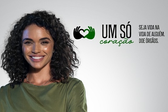 A imagem mostra uma mulher jovem com um grande sorriso, vestindo uma blusa verde oliva. Ela tem cabelos encaracolados e pretos e parece estar muito feliz. Ao lado dela, há um logotipo e um slogan que diz "Um só coração" com um ícone de dois corações entrelaçados e a mensagem "Seja vida na vida de alguém. Doe órgãos." A composição sugere que a imagem faz parte de uma campanha de incentivo à doação de órgãos, promovendo uma mensagem positiva e encorajadora sobre o impacto pessoal que alguém pode ter ao doar órgãos. O fundo é neutro e claro, destacando o rosto sorridente da mulher e a mensagem da campanha.