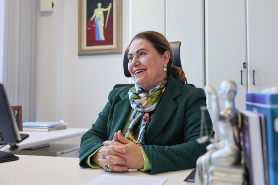 A imagem mostra uma mulher sentada em uma cadeira de escritório, sorrindo. Ela está usando um blazer verde e um lenço colorido ao redor do pescoço. Suas mãos estão cruzadas sobre a mesa à sua frente. No fundo, há um armário branco e uma pintura na parede que parece representar a deusa da justiça, com uma balança em uma das mãos. Sobre a mesa, há alguns objetos, incluindo uma estatueta prateada e alguns livros ou pastas empilhados. A atmosfera parece ser de um ambiente de trabalho, possivelmente um escritório.