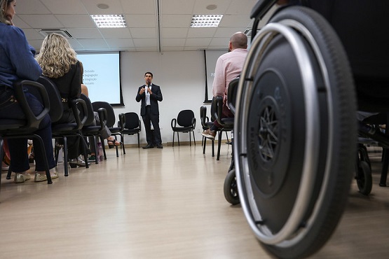 A imagem mostra uma sala de conferências ou reunião, onde um homem de terno está em pé, falando para um grupo de pessoas sentadas em cadeiras. Ele está de frente para a audiência, que está disposta em duas fileiras, uma de cada lado, deixando um corredor no meio. No canto direito da imagem, em primeiro plano, há uma roda de cadeira de rodas, indicando que há uma pessoa em cadeira de rodas presente na reunião. O ambiente é bem iluminado, com luzes fluorescentes no teto, e há uma tela de projeção ao fundo, embora o conteúdo projetado não seja legível na imagem.