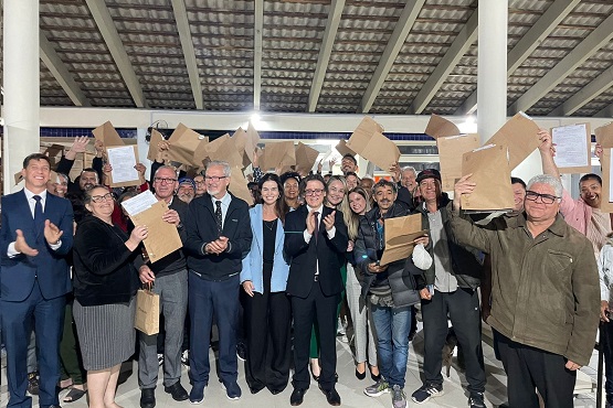 A imagem mostra um grupo grande de pessoas reunidas em um espaço coberto. A maioria delas está segurando envelopes marrons ou documentos, levantando-os no ar com expressões de felicidade e celebração. No centro do grupo, há algumas pessoas vestidas formalmente, como se fossem autoridades ou organizadores do evento. O ambiente parece ser um local público ou uma área de eventos, com um teto alto e colunas brancas. As pessoas estão sorrindo e algumas estão aplaudindo.
