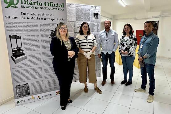 A imagem mostra cinco pessoas em pé, lado a lado, em frente a um grande painel que parece ser uma exposição. O painel tem o título "Diário Oficial Estado de Santa Catarina" e menciona "Do prelo ao digital: 90 anos de história e transparência". As pessoas estão em um ambiente interno com piso de azulejo branco e paredes claras. A primeira pessoa à esquerda é uma mulher de cabelos loiros, usando óculos e vestindo roupas escuras. Ao lado dela, há outra mulher de cabelos castanhos, usando óculos e uma blusa listrada. No centro, um homem de barba, vestindo uma camisa clara e calça jeans. A quarta pessoa é uma mulher de cabelos castanhos, usando uma blusa com estampas. Por último, à direita, está um homem de cabelos curtos, vestindo uma camisa jeans e calça escura. Todos estão sorrindo e olhando para a câmera.