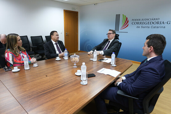 A imagem mostra uma reunião em uma sala de conferências. Quatro pessoas estão sentadas ao redor de uma mesa de madeira. À esquerda, uma mulher com cabelo comprido veste uma blusa colorida e está ao lado de um homem de terno escuro. Do outro lado da mesa, há dois homens de terno, um deles usando óculos. Na mesa, há garrafas de água, xícaras de café e um recipiente de vidro com o que parece ser balas ou pequenos doces. Na parede ao fundo, há um logotipo e o texto "Poder Judiciário de Santa Catarina" “Corregedoria-Geral”. A sala tem um ambiente formal e corporativo.