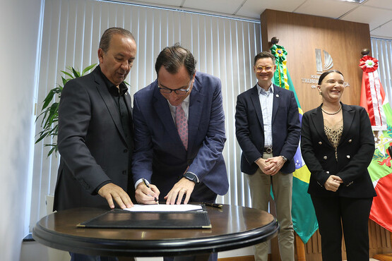 A imagem mostra quatro pessoas em um ambiente formal, provavelmente um escritório ou sala de reuniões. No centro, um homem de terno azul e gravata rosa está assinando um documento sobre uma mesa redonda. Ao seu lado esquerdo, outro homem de terno preto observa a assinatura. Atrás deles, há mais duas pessoas: um homem de terno azul escuro e calça bege, e uma mulher de terno preto, ambos sorrindo. No fundo, há uma parede com persianas brancas e duas bandeiras, uma do Brasil e outra de Santa Catarina, decoradas com laços. A parede também possui um logotipo ou emblema, mas não é possível identificar os detalhes.