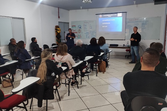 A imagem mostra uma sala de aula ou de conferências com várias pessoas sentadas em cadeiras com pranchetas, voltadas para a frente da sala. Na frente, há duas pessoas em pé, uma delas está falando e a outra está ao lado, possivelmente aguardando sua vez de falar. Na parede da frente, há um projetor exibindo uma apresentação em um telão. O slide projetado tem um fundo azul e parece conter texto, mas não é possível ler o conteúdo específico. As paredes da sala são brancas e há um quadro branco ao lado do telão. As pessoas na sala estão prestando atenção na apresentação.