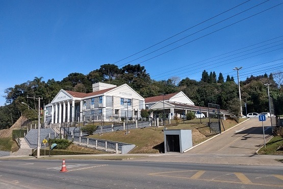 A imagem mostra um prédio de arquitetura clássica, com colunas na fachada, que parece ser um fórum, pois há uma placa com a palavra "FÓRUM" no topo. O edifício está localizado em uma área elevada, com escadas e rampas de acesso. Há uma rua em frente ao prédio, com uma faixa de pedestres e um cone de trânsito laranja. Ao fundo, há árvores e vegetação densa, sugerindo que o local está em uma área arborizada. O céu está claro e azul, indicando um dia ensolarado.