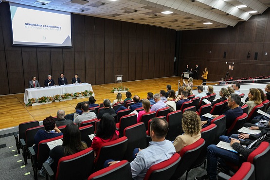 A imagem mostra um auditório com várias pessoas sentadas em cadeiras vermelhas, assistindo a um evento. Na frente do auditório, há uma mesa comprida coberta com uma toalha branca, onde estão sentadas quatro pessoas, provavelmente palestrantes ou membros de uma mesa de discussão. Atrás deles, há um grande telão projetando a imagem de um slide que diz "SEMINÁRIO CATARINENSE". O ambiente é bem iluminado e as paredes são revestidas de madeira escura. Algumas pessoas estão tomando notas enquanto assistem.