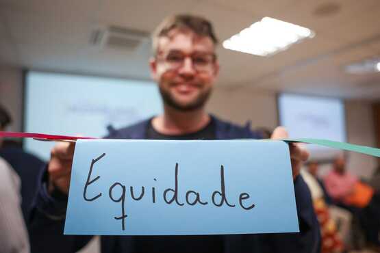 A imagem mostra um homem sorridente segurando um cartão azul com a palavra "Equidade" escrita nele. Ele está em um ambiente interno, possivelmente uma sala de conferências ou reunião, com outras pessoas ao fundo. A iluminação é clara, com luzes de teto visíveis. O homem usa óculos e tem barba curta. A cena parece ser parte de um evento ou workshop relacionado a temas de inclusão ou justiça social.