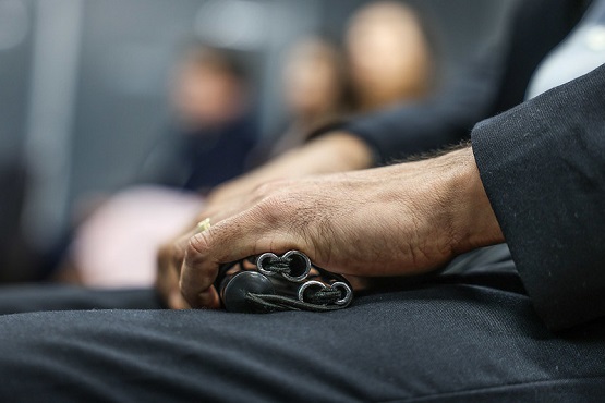 A imagem mostra uma mão masculina segurando uma bengala dobrada. A mão está descansando sobre uma perna, que está coberta por uma calça preta. A pessoa está vestindo um terno ou blazer preto. O fundo da imagem está desfocado, mas parece haver outras pessoas sentadas, sugerindo que a cena pode ser em um ambiente como uma sala de espera ou uma reunião.
