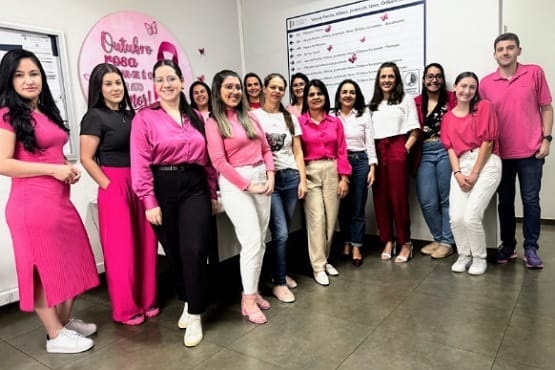 A imagem mostra um grupo de 14 pessoas, a maioria mulheres, posando para uma foto em um ambiente interno. A maioria delas está vestindo roupas em tons de rosa, o que sugere que estão participando de uma campanha de conscientização do Outubro Rosa, que é um movimento internacional de conscientização para o controle do câncer de mama. No fundo, há um cartaz circular na parede com a inscrição "Outubro Rosa". As pessoas estão sorrindo e parecem estar em um ambiente de trabalho ou evento relacionado ao tema.