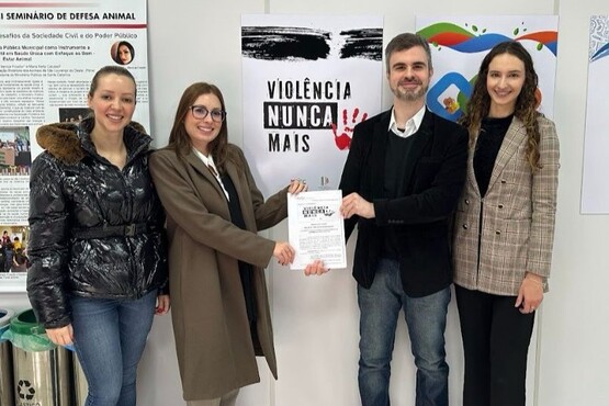 A imagem mostra quatro pessoas posando para uma foto em frente a um cartaz que diz "Violência Nunca Mais". As duas pessoas no centro estão segurando um documento. A pessoa à esquerda está vestindo uma jaqueta preta acolchoada e jeans, a segunda pessoa está usando um casaco marrom e óculos, a terceira pessoa, um homem, está vestindo um blazer preto sobre uma camisa branca e jeans, e a pessoa à direita está usando um blazer xadrez e calças escuras.