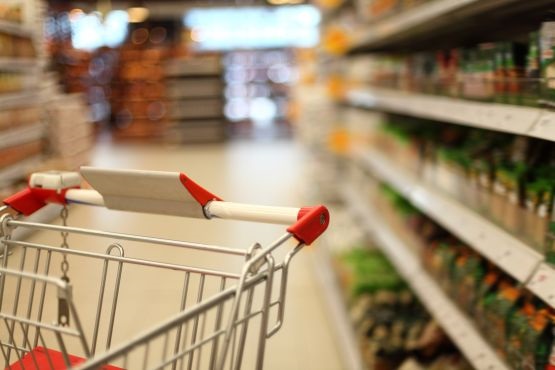 A imagem mostra o interior de um supermercado. Em primeiro plano, há um carrinho de compras com detalhes em vermelho nas alças. O carrinho está vazio e posicionado no corredor entre duas prateleiras. À direita, as prateleiras estão cheias de produtos, mas estão desfocadas, dificultando a identificação dos itens. Ao fundo, a imagem também está desfocada, mostrando mais prateleiras e produtos. A iluminação é clara, típica de um ambiente de supermercado.