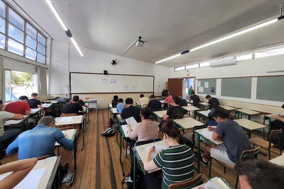 A imagem mostra uma sala de aula com várias pessoas sentadas em carteiras, aparentemente fazendo uma prova ou atividade escrita. A sala é ampla, com piso de madeira e janelas grandes à esquerda, permitindo a entrada de luz natural. As carteiras estão organizadas em fileiras, e as pessoas estão concentradas em seus papeis. Na frente da sala, há um quadro branco com algumas anotações. O ambiente parece ser de um exame ou teste, dada a postura dos alunos e a disposição das carteiras.