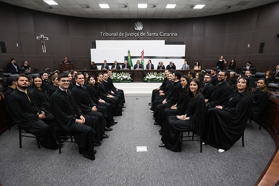 A imagem mostra um grupo de pessoas, vestidas com togas pretas, sentadas em um auditório. Elas parecem ser advogados ou juízes. No fundo da sala, há um painel com o texto "Tribunal de Justiça de Santa Catarina" e uma bandeira do Brasil ao centro. Há também um crucifixo pendurado acima do painel. A disposição dos assentos e a decoração formal sugerem que este é um evento solene ou uma cerimônia oficial. As pessoas na imagem exibem expressões sérias e atentas, típicas de um ambiente judicial ou de uma ocasião formal.