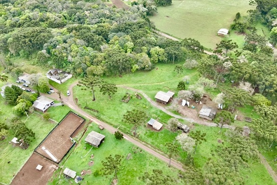 A imagem mostra uma vista aérea de uma paisagem rural. Há várias casas pequenas e construções espalhadas, cercadas por áreas verdes. Estradas de terra conectam as diversas propriedades. Em algumas partes, pode-se ver terrenos com vegetação mais densa, possivelmente pequenas áreas de floresta. Além disso, há campos abertos que parecem ser usados para agricultura ou pastagem. A área geral apresenta uma mistura de uso residencial e agrícola, típica de uma zona rural.