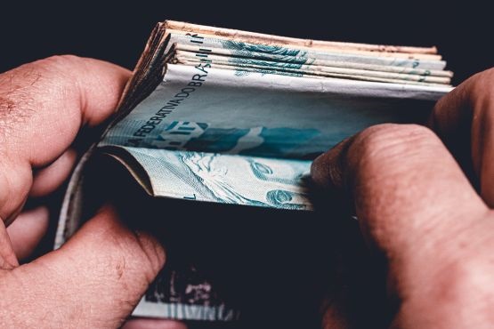 A imagem mostra as mãos de uma pessoa segurando uma quantidade significativa de notas de dinheiro dobradas. As notas parecem ser de real brasileiro, com a cor predominante azul, que é típica das notas de cinquenta reais. O foco está no topo das notas, tornando os detalhes nas extremidades um pouco menos nítidos. O fundo é escuro, destacando ainda mais o dinheiro nas mãos da pessoa. As mãos parecem ser de um adulto, com detalhes bem visíveis, como as linhas e texturas da pele.