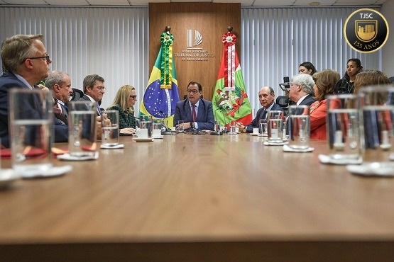 A imagem mostra uma reunião formal com várias pessoas sentadas ao redor de uma longa mesa retangular. No centro da mesa, há duas bandeiras, uma do Brasil e outra de Santa Catarina. Todos os participantes parecem estar envolvidos em uma discussão séria ou apresentação, voltados para a pessoa sentada no centro, que parece liderar a reunião. Há copos de água diante de cada participante, sugerindo uma configuração típica de uma reunião de negócios ou governamental. A sala é bem iluminada e tem um aspecto formal, com decoração minimalista.