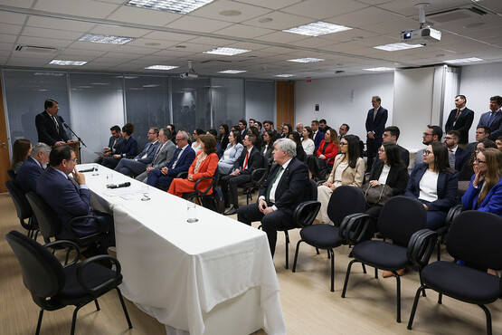 A imagem mostra um ambiente corporativo onde várias pessoas estão reunidas em uma sala de conferência. Há cerca de vinte pessoas sentadas e algumas em pé, todas voltadas para um homem que está de pé falando no centro da sala. O público é composto por homens e mulheres vestidos formalmente. A sala tem um design moderno, com cadeiras acolchoadas, uma longa mesa com um pano branco na frente e um quadro branco ao fundo.