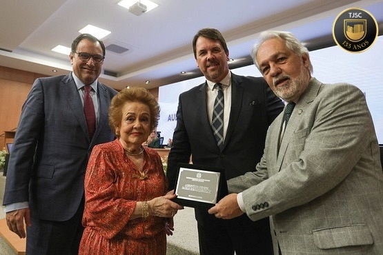 A imagem mostra quatro pessoas em um ambiente formal, provavelmente em um evento ou cerimônia. À esquerda, há um homem de óculos usando um terno azul e gravata vermelha. Ao lado dele, está uma mulher idosa com cabelo castanho claro, usando um vestido vermelho com detalhes dourados. No centro, há um homem de terno preto e gravata listrada. À direita, outro homem de cabelo grisalho e barba, usando um terno cinza, está segurando uma placa ou prêmio junto com a mulher. Ao fundo, há um auditório com um telão exibindo parte de um texto que não está completamente visível. A cena parece ser de uma entrega de prêmio ou homenagem.