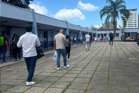 A imagem mostra uma fila de pessoas em um ambiente externo, possivelmente em frente a um prédio público ou escola. As pessoas estão alinhadas em um pátio pavimentado com blocos de concreto, aguardando para entrar em uma área coberta à esquerda. Algumas pessoas estão vestindo roupas casuais, como calças jeans e camisetas, e carregam bolsas ou mochilas. Ao fundo, há uma palmeira alta e um céu azul com algumas nuvens. No lado direito, é possível ver um edifício alto ao longe.