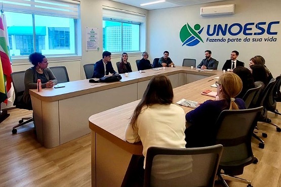 A imagem mostra uma sala de reuniões com uma mesa em formato de "U" ao redor da qual estão sentadas várias pessoas. Há um total de doze pessoas na sala, incluindo homens e mulheres, todos vestidos de forma formal ou semi-formal. Na parede ao fundo, há um logotipo grande com o texto "UNOESC Fazendo parte da sua vida". A sala é bem iluminada, com janelas ao fundo que deixam entrar luz natural. No canto esquerdo da imagem, há uma bandeira, possivelmente do estado de Santa Catarina. A atmosfera parece ser de uma reunião ou discussão formal.