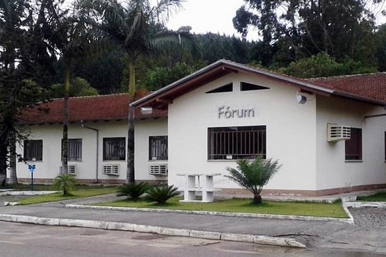 A imagem mostra um edifício de um andar com um telhado de duas águas coberto por telhas vermelhas. Na fachada do prédio, há a palavra "Fórum" escrita em letras grandes. O edifício possui várias janelas com grades e aparelhos de ar-condicionado instalados. Em frente ao prédio, há uma área gramada com algumas plantas e palmeiras. O local parece estar em uma área arborizada, com várias árvores ao fundo.