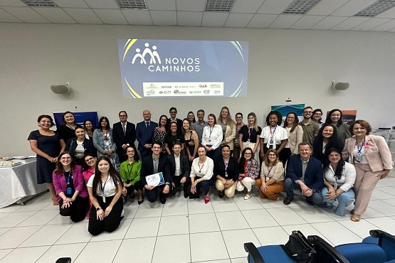 A imagem mostra um grupo de aproximadamente trinta pessoas posando para uma foto em um ambiente interno, que parece ser uma sala de conferência ou de aula. Ao fundo, há um grande banner com o logotipo e o nome "Novos Caminhos", e o subtítulo "Eixo de ação das redes estaduais". As pessoas no grupo são diversas, incluindo homens e mulheres de diferentes idades e etnias, vestindo roupas formais e semi-formais. Eles estão sorrindo e parecem estar participando de um evento ou workshop relacionado ao programa "Novos Caminhos".
