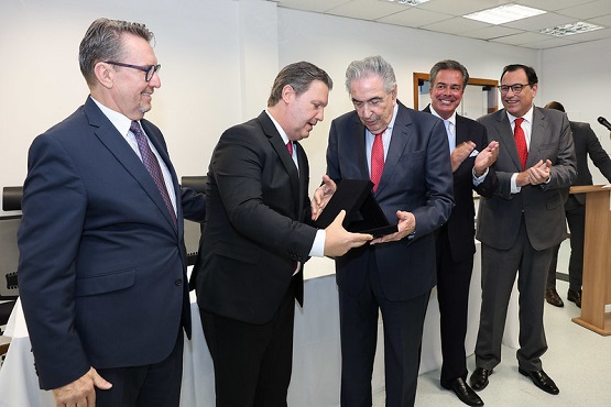 A imagem mostra um grupo de cinco homens em um ambiente interno, provavelmente durante um evento formal ou cerimônia. Os homens estão vestidos formalmente em ternos e gravatas. O homem ao centro está recebendo ou mostrando um objeto pequeno, que parece ser uma placa ou um prêmio, a outro homem que o observa atentamente. Os outros três homens ao redor estão sorrindo e aplaudindo, sugerindo um momento de celebração ou reconhecimento. O ambiente tem um aspecto de escritório ou sala de conferências, com paredes claras e um ambiente bem iluminado.
