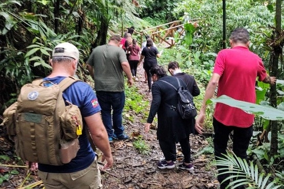 A imagem mostra um grupo de pessoas em um campo aberto, cercado por uma floresta densa. Ao fundo, pode-se ver uma cascata alta, descendo pelas montanhas cobertas de vegetação verde exuberante. O grupo parece estar envolvido em alguma atividade ao ar livre, possivelmente um passeio ou uma aula de campo, dado que algumas pessoas estão manuseando equipamentos como câmeras e guarda-chuvas, enquanto outras estão conversando e apontando para a natureza ao redor. O cenário é bastante pitoresco e transmite uma sensação de tranquilidade e conexão com a natureza.