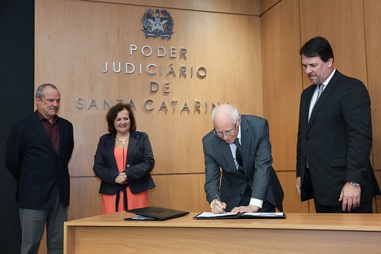 A imagem mostra quatro pessoas em uma sala de reuniões oficial com um painel de madeira ao fundo onde se lê "PODER JUDICIÁRIO DE SANTA CATARINA". Três homens e uma mulher, todos vestidos formalmente. Um dos homens está assinando um documento sobre uma mesa, enquanto os outros observam. A mulher e os dois outros homens parecem estar em um momento de solenidade ou de observação do ato de assinatura. Todos parecem sérios e concentrados na tarefa que está sendo executada.