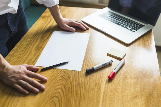 A imagem mostra uma pessoa em uma mesa de trabalho organizada com diversos itens. À vista estão um laptop aberto, um papel em branco, um par de marcadores, e um pequeno bloco de notas. A pessoa está com as mãos sobre a mesa, aparentemente em posição de começar a trabalhar ou organizar seus pensamentos. A cena sugere um ambiente de escritório ou de estudo, onde a preparação para uma tarefa criativa ou administrativa está prestes a acontecer. A iluminação suave e o fundo desfocado ajudam a focar a atenção nos elementos sobre a mesa.
