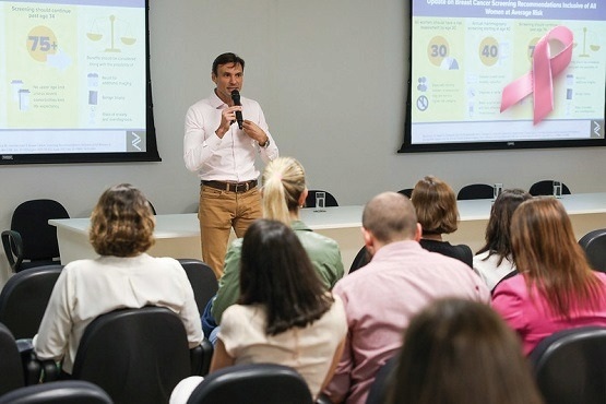 A imagem mostra uma cena de uma palestra ou seminário em uma sala de conferência. Um homem está em pé na frente, falando ao microfone e segurando uma caneta, aparentemente explicando algo importante. Ele está vestido de forma casual-profissional com camisa de manga longa e calça clara. Atrás dele, há um grande telão mostrando slides com estatísticas e informações sobre o câncer de mama, indicado por uma faixa rosa no canto. O público, formado por várias pessoas sentadas em cadeiras, parece atento ao que está sendo dito. A configuração sugere que o tema da apresentação é relacionado à saúde, possivelmente um treinamento ou informação sobre recomendações de rastreamento de câncer.