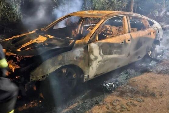 A imagem mostra um carro bastante danificado e queimado, aparentemente após um incêndio. O veículo está sem as janelas, com a pintura totalmente destruída, revelando a lataria enegrecida e deformada pelo fogo. Há fumaça saindo dos restos do carro, indicando que o incêndio pode ter sido recente. Ao fundo, percebe-se uma área que parece ser um terreno aberto ou uma estrada de terra. Um bombeiro, parcialmente visível no lado esquerdo da imagem, está próximo ao carro, sugerindo uma cena de resgate ou contenção de incêndio.