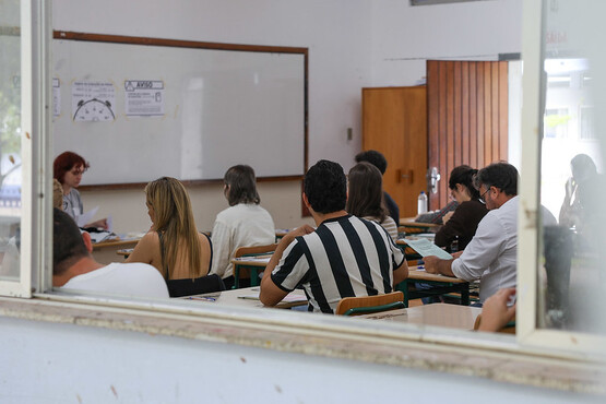 A imagem mostra uma sala de aula com adultos assistindo a uma aula. Eles estão sentados em carteiras escolares enfileiradas, voltados para uma mulher ruiva que está à frente da classe, perto de uma lousa branca. A mulher parece estar explicando algo, segurando papéis em uma das mãos. A sala tem paredes brancas, uma porta de madeira aberta ao fundo, e uma lousa que contém escritas e desenhos parcialmente visíveis. A cena é vista de fora da sala, através de uma janela, o que sugere a perspectiva de alguém observando a aula de fora.