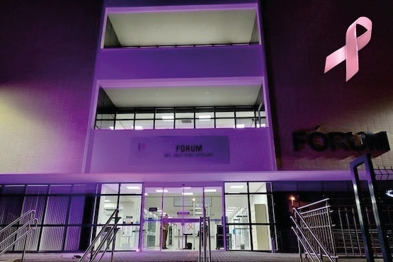 A imagem mostra a fachada de um prédio identificado como "Fórum", iluminado com luzes roxas. O prédio tem três andares visíveis, com varandas nos dois andares superiores. Na parte inferior, há uma entrada com portas de vidro e corrimãos nas escadas que levam até ela. No canto superior direito da imagem, há um laço rosa, que é um símbolo frequentemente associado à conscientização sobre o câncer de mama. Na parede acima da entrada, há uma placa com o texto "FÓRUM" e abaixo, em letras menores, "DR. JOSÉ FERNANDES VITURINO".
