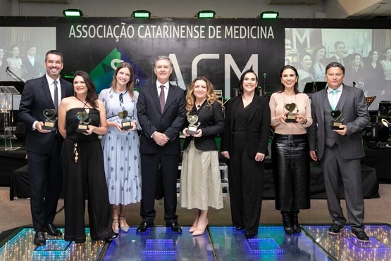 A imagem retrata um grupo de sete pessoas posando para uma fotografia em um evento formal, aparentemente uma cerimônia de premiação. Cada pessoa segura um troféu, sugerindo que receberam algum tipo de reconhecimento ou prêmio. O evento parece ser organizado pela Associação Catarinense de Medicina, como indicado pelo logo e pelo nome visível ao fundo em um grande banner. Os indivíduos estão vestidos formalmente—os homens com ternos e gravatas, e as mulheres com vestidos e saias elegantes. A cenografia inclui iluminação sofisticada e um piso que reflete luz, o que adiciona um brilho especial à ocasião. Eles parecem estar em um palco, com luzes embutidas no chão que iluminam o espaço de forma dramática, realçando a importância do momento capturado.