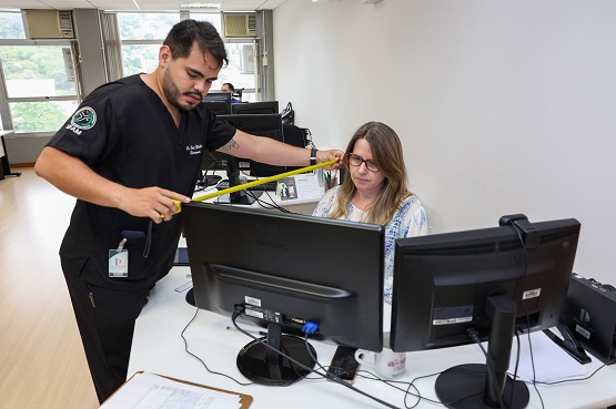 A imagem mostra um homem e uma mulher em um ambiente de escritório trabalhando juntos em um computador. O homem, vestindo um uniforme preto com emblemas, está medindo algo com uma fita métrica amarela, talvez ajustando ou verificando o espaço para ergonomia ou algum outro propósito relacionado à manutenção ou segurança do local de trabalho. A mulher, sentada e vestindo óculos e um suéter claro, observa o monitor enquanto aparentemente discute o processo ou resultado com o colega. O cenário inclui duas grandes telas de computador, documentos e outros itens de escritório sobre a mesa, indicando um ambiente profissional ativo e funcional. As janelas ao fundo permitem a entrada de luz natural, contribuindo para uma atmosfera clara e aberta no escritório.