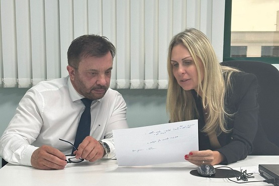 A imagem mostra um homem e uma mulher, ambos vestidos formalmente, analisando um documento em uma mesa de escritório. O homem, de camisa branca e gravata, segura uma caneta, indicando que ele está pronto para escrever ou assinar o documento. A mulher, usando um blazer escuro e blusa clara, segura o papel e aponta para algo específico, possivelmente discutindo ou explicando um ponto importante do documento. Eles estão sentados lado a lado em frente a uma grande janela com cortinas verticais, que permite a entrada de luz natural, mas mantém a privacidade do ambiente. Há também objetos como óculos e outros papéis sobre a mesa, o que sugere que estão em um ambiente de trabalho, possivelmente um escritório ou sala de reuniões. A interação entre os dois parece séria e focada, típica de um contexto profissional onde decisões importantes estão sendo consideradas ou acordos estão sendo finalizados.