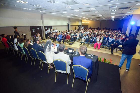 A imagem mostra um evento em um grande auditório. Há uma multidão de pessoas sentadas em cadeiras dispostas em frente a um palco, onde uma apresentação parece estar ocorrendo. Na frente da plateia, há uma mesa com seis pessoas sentadas, possivelmente juízes ou palestrantes, voltadas para a multidão. Elas estão ouvindo atentamente o que está sendo dito. A sala é ampla e bem iluminada, e a atmosfera sugere um evento formal, como uma conferência ou semi