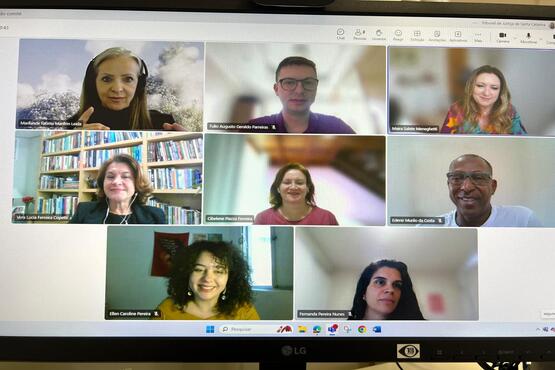 A imagem exibe a tela de um computador durante uma reunião virtual com oito participantes, cada um mostrado em sua própria janela. Cada participante parece estar em um ambiente diferente, variando de escritórios com prateleiras de livros a ambientes mais casuais em casa. Os participantes têm diferentes expressões, mas todos parecem estar atentos à discussão. A configuração sugere uma reunião de trabalho ou um webinar, com pessoas de diferentes backgrounds participando de uma conversa ou apresentação online.