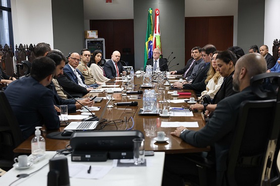 A imagem mostra uma reunião formal em uma sala de conferências. Há várias pessoas sentadas ao redor de uma mesa retangular de madeira. Na mesa, há garrafas de água, copos, papéis e alguns laptops. Ao fundo, há duas bandeiras, uma do Brasil e outra de um estado ou instituição, que não é possível identificar claramente. As pessoas na reunião estão vestidas formalmente, com ternos e roupas sociais. A sala tem paredes brancas e portas de madeira. Parece ser uma reunião de negócios ou governamental, dada a formalidade do ambiente e das vestimentas.