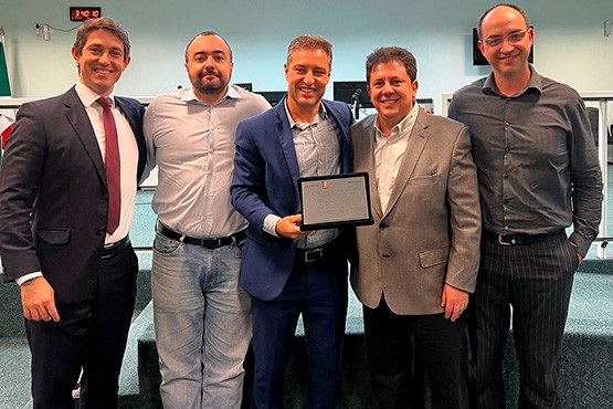 A imagem mostra cinco homens posando juntos para uma foto em um ambiente interno, que parece ser uma sala de conferências ou uma câmara. Todos estão sorrindo. O homem no centro está segurando uma placa ou certificado. Ele veste um terno azul escuro com uma camisa azul clara. À sua esquerda, há um homem de camisa branca e calça jeans, e à sua direita, um homem de terno cinza claro. Os dois homens nas extremidades estão usando ternos; o da esquerda veste um terno escuro com gravata vermelha, e o da direita veste um terno escuro com uma camisa cinza. Ao fundo, há uma parede clara e um painel eletrônico com números vermelhos