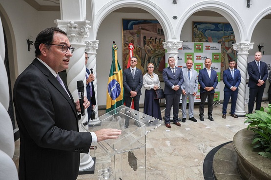 A imagem mostra um evento formal em um ambiente interno com arquitetura clássica, com colunas brancas e arcos. À esquerda, um homem de terno escuro e óculos está falando ao microfone em um púlpito transparente. Ao fundo, há várias pessoas de pé, todas vestidas formalmente, incluindo homens de terno e mulheres de saia e blusa. Atrás delas, há uma bandeira do Brasil e um painel com logotipos. O ambiente parece ser um salão com decoração elegante, incluindo um mural na parede.