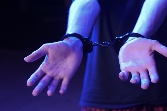 A imagem mostra um par de mãos algemadas. As algemas são de metal e estão prendendo os pulsos de uma pessoa. A pessoa está usando uma camiseta preta e a imagem tem uma iluminação azulada, destacando as mãos e as algemas. As mãos estão voltadas para cima, com os dedos ligeiramente abertos.
