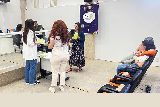 A imagem mostra uma sala de aula ou conferência com várias pessoas. No centro, há três mulheres em pé, conversando entre si. Uma delas está de costas, com cabelo cacheado e vestindo uma roupa clara. As outras duas estão de frente, uma com cabelo liso e longo, usando uma camisa branca e calça jeans, e a outra com cabelo preso, usando um vestido estampado e um casaco azul. Ao fundo, há uma mesa com duas pessoas sentadas, aparentemente observando ou participando da conversa. À direita, há duas pessoas sentadas em cadeiras acolchoadas, uma delas com a mão no queixo, parecendo atenta. Na parede ao fundo, há um cartaz roxo com o texto "CPAM/S" e "PISC", que parece ser parte de um evento ou programa. A sala tem um ambiente formal, possivelmente em uma instituição educacional ou governamental.