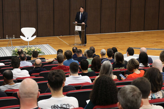 A imagem mostra uma palestra ou conferência em um auditório. No centro da imagem, há um homem de terno escuro e gravata, em pé atrás de um púlpito transparente, falando para o público. À esquerda do palestrante, há uma cadeira branca moderna e um arranjo de flores sobre um tapete listrado. O público está sentado em cadeiras vermelhas, e a maioria das pessoas está de costas para a câmera, olhando para o palestrante. O chão é de madeira clara, e a parede ao fundo é revestida de painéis de madeira escura.