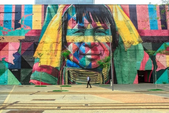 A imagem mostra um mural colorido pintado em uma grande parede. O mural retrata o rosto de uma pessoa indígena com um sorriso suave. Ela está usando um adorno de pescoço tradicional. O rosto é composto por várias cores vibrantes, como amarelo, azul, rosa e verde, que se misturam em padrões geométricos. Em frente ao mural, há uma calçada e uma pessoa caminhando, o que dá uma ideia da escala da pintura. A arte é rica em detalhes e cores, criando um impacto visual impressionante.
