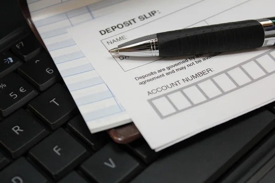 A imagem mostra um teclado de computador com um formulário de depósito bancário ("Deposit Slip") colocado sobre ele, juntamente com uma caneta preta e prateada repousando sobre o formulário. O formulário tem campos típicos, como "Name" (nome) e "Account Number" (número da conta), com espaço para preenchimento. Ao lado do formulário de depósito, também é visível uma folha com linhas, possivelmente de um talão de cheques ou um caderno de anotações. A cena sugere uma situação de trabalho ou transação financeira, onde a pessoa pode estar preenchendo um depósito ou organizando documentos bancários.