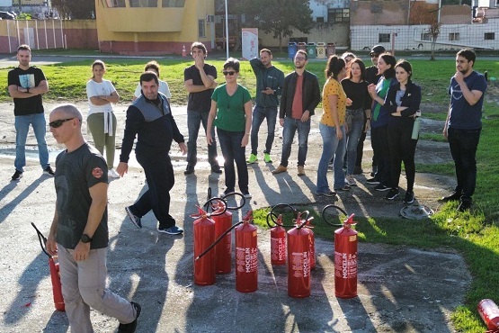 A imagem mostra um grupo de pessoas reunidas ao ar livre, aparentemente participando de um treinamento de combate a incêndio. No centro da cena, há vários extintores de incêndio vermelhos posicionados no chão, alinhados e prontos para uso. À esquerda, um homem com óculos de sol e camiseta preta está caminhando, provavelmente um instrutor ou responsável pelo treinamento. Ao fundo, o grupo de pessoas, composto por homens e mulheres, está observando atentamente. Algumas pessoas estão conversando entre si, enquanto outras parecem prestes a participar da atividade. O cenário ao redor inclui uma área aberta com grama e edifícios ao fundo, sugerindo que o treinamento está ocorrendo em uma instituição ou espaço público.